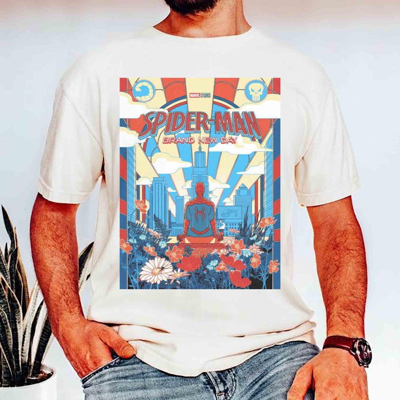 May include: A cream-colored t-shirt featuring a graphic print of Spider-Man sitting in a meditative pose. The design includes a city skyline, clouds, and colorful flowers in red, blue, and white, with the text Spider-Man Brand New Day.