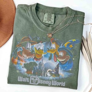 K&ouml;nnte beinhalten: Olivgr&uuml;nes T-Shirt mit einem Disney-Motiv, das Micky Maus, Donald Duck, Goofy und Pluto vor dem Cinderella-Schloss zeigt. Der Text "Walt Disney World" ist darunter aufgedruckt. Eine silberne Halskette mit Anh&auml;nger liegt auf dem Shirt.