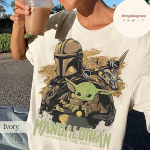 Puede incluir: Camiseta color marfil con un dise&ntilde;o de The Mandalorian y Grogu, con una nave espacial de fondo. El texto "MANDALORIAN - THIS IS THE WAY" est&aacute; impreso debajo. La camiseta es de color claro.