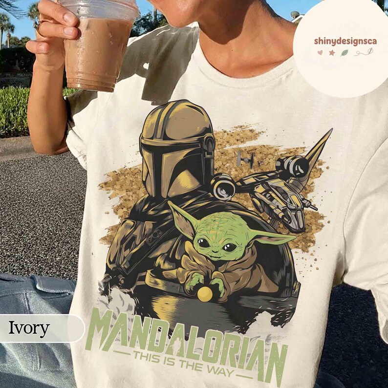 Puede incluir: Camiseta color marfil con un dise&ntilde;o de The Mandalorian y Grogu, con una nave espacial de fondo. El texto "MANDALORIAN - THIS IS THE WAY" est&aacute; impreso debajo. La camiseta es de color claro.
