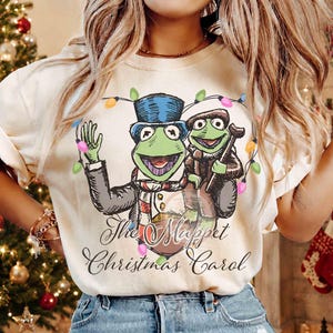 May include: A cream-colored t-shirt featuring a graphic illustration of two green frog characters, one wearing a blue top hat and the other a brown cap, surrounded by colorful Christmas lights with the text The Muppet Christmas Carol.