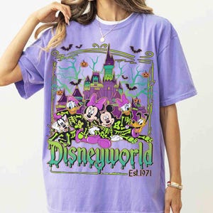 May include: A lilac t-shirt with a Disney World Halloween design. The graphic shows Mickey and Minnie Mouse, Donald and Daisy Duck, Goofy, and Pluto in skeleton costumes, with a castle, bats, and pumpkins. The text "Disneyworld Est. 1971" is also included.