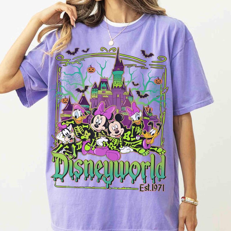 May include: A lilac t-shirt with a Disney World Halloween design. The graphic shows Mickey and Minnie Mouse, Donald and Daisy Duck, Goofy, and Pluto in skeleton costumes, with a castle, bats, and pumpkins. The text "Disneyworld Est. 1971" is also included.