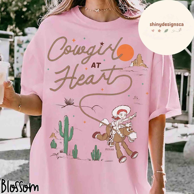 May include: Pink t-shirt with the text "Cowgirl at Heart" in a rope-style font. The graphic features a cartoon cowgirl riding a horse, with a desert landscape and a setting sun. The shirt has a relaxed fit.