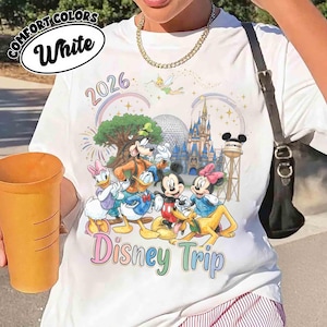 May include: A white t-shirt featuring a colorful graphic of Disney characters including Mickey Mouse, Minnie Mouse, Donald Duck, Daisy Duck, Goofy, and Pluto. The design includes the text 2026 and Disney Trip, along with a castle and Tinker Bell.