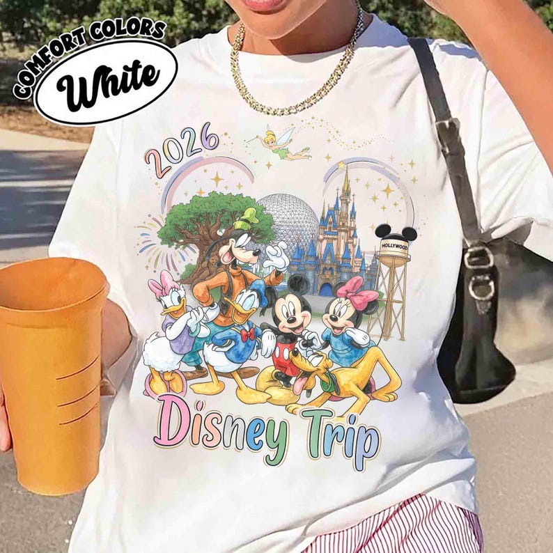May include: A white t-shirt featuring a colorful graphic of Disney characters including Mickey Mouse, Minnie Mouse, Donald Duck, Daisy Duck, Goofy, and Pluto. The design includes the text 2026 and Disney Trip, along with a castle and Tinker Bell.
