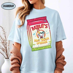 Puede incluir: Camiseta azul claro con un gr&aacute;fico de una bolsa de snacks con el texto "MAUI'S BOAT SNACK UPGRADE". El dise&ntilde;o incluye personajes de dibujos animados y una paleta de colores vibrantes, ideal para fans de pel&iacute;culas animadas.