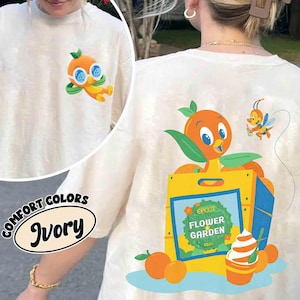 Puede incluir: Camiseta de color marfil con un diseño de pájaro naranja de dibujos animados. La parte trasera presenta un gráfico más grande del pájaro en una caja amarilla, con el texto "EPCOT INTERNATIONAL FLOWER GARDEN FESTIVAL 2024". También hay una abeja de dibujos animados.