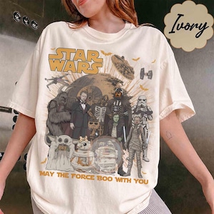 May include: An ivory t-shirt featuring a Star Wars graphic with characters in Halloween costumes, including a ghost, skeleton, and mummy. The text reads Star Wars and May The Force Boo With You, surrounded by bats and a Death Star illustration.