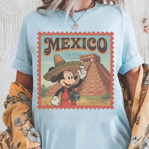 May include: Pale blue t-shirt with a retro graphic. It shows Mickey Mouse in a sombrero, holding a camera, with the word "MEXICO" above a pyramid illustration. The design is framed within a postage stamp border.