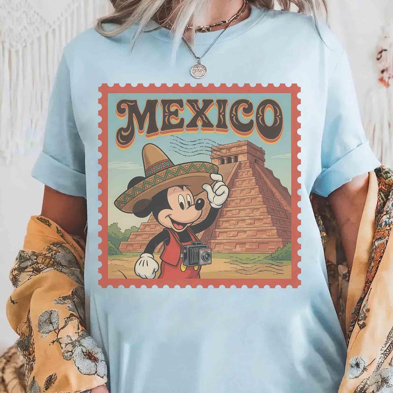 May include: Pale blue t-shirt with a retro graphic. It shows Mickey Mouse in a sombrero, holding a camera, with the word "MEXICO" above a pyramid illustration. The design is framed within a postage stamp border.
