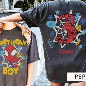 Pu&ograve; includere: Una t-shirt grigio antracite con una stampa grafica di un supereroe rosso e blu sul davanti con il testo "BIRTHDAY BOY" e "web-out!". Il retro mostra un grande numero 3 rosso con motivo a ragnatela, stelle e la scritta "Name".