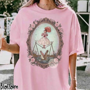 Puede incluir: Una camiseta de color rosa claro con un gr&aacute;fico central de una persona con vestido sosteniendo un paraguas mientras camina sobre una cuerda floja encima de un caim&aacute;n. La ilustraci&oacute;n est&aacute; en un marco ovalado adornado con rosas.