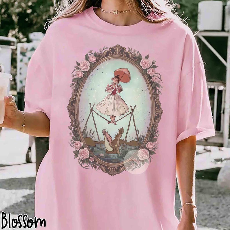 Puede incluir: Una camiseta de color rosa claro con un gr&aacute;fico central de una persona con vestido sosteniendo un paraguas mientras camina sobre una cuerda floja encima de un caim&aacute;n. La ilustraci&oacute;n est&aacute; en un marco ovalado adornado con rosas.
