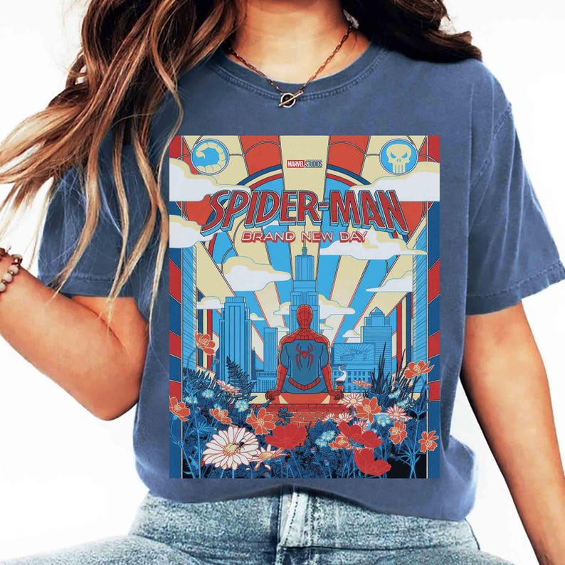 May include: A blue t-shirt featuring a graphic print of Spider-Man sitting before a city skyline. The design includes the text "SPIDER-MAN BRAND NEW DAY" and "MARVEL STUDIOS," surrounded by red, blue, and yellow flowers and clouds.