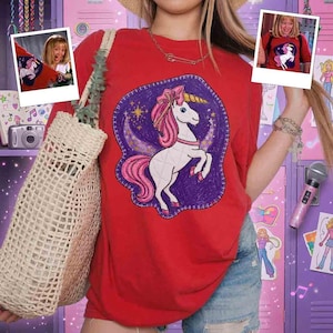 May include: A bright red short-sleeve t-shirt featuring a central graphic of a white unicorn with a pink mane and tail, set against a purple crescent moon and star background. The shirt is paired with a woven beige tote bag and blue denim shorts.