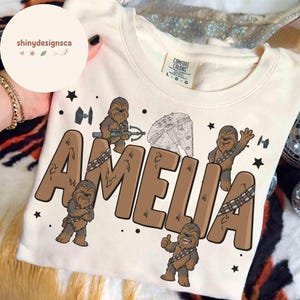 Può includere: Felpa color crema con il nome "AMELIA" in lettere marroni, illustrazioni di Chewbacca e grafica di Star Wars. La maglietta ha un'etichetta Comfort Colors.