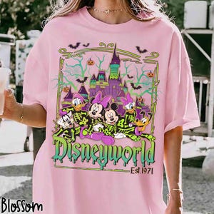 May include: Pale pink t-shirt with a Disneyworld graphic. The design features Mickey, Minnie, Donald, Daisy, and Goofy in skeleton costumes, a castle, bats, and the text "Disneyworld Est. 1971".