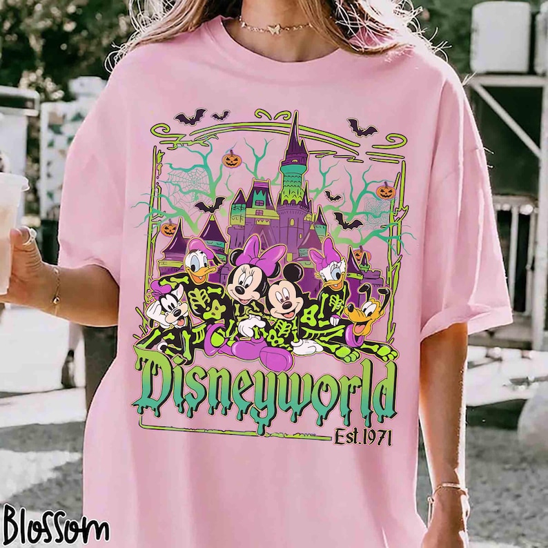 May include: Pale pink t-shirt with a Disneyworld graphic. The design features Mickey, Minnie, Donald, Daisy, and Goofy in skeleton costumes, a castle, bats, and the text "Disneyworld Est. 1971".