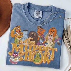 Może przedstawiać: Niebieska koszulka Comfort Colors z grafiką postaci z Muppet Show. Koszulka ma okrągły dekolt i krótkie rękawy. Logo Muppet Show jest pomarańczowe i żółte. Złoty naszyjnik jest zarzucony na koszulkę.