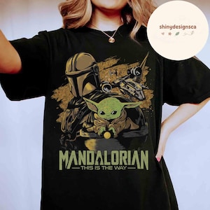 Puede incluir: Camiseta negra con un gr&aacute;fico de The Mandalorian y Grogu, una nave espacial y el texto "MANDALORIAN - THIS IS THE WAY" en verde y dorado. El dise&ntilde;o incluye una figura con casco y un personaje alien&iacute;gena verde.
