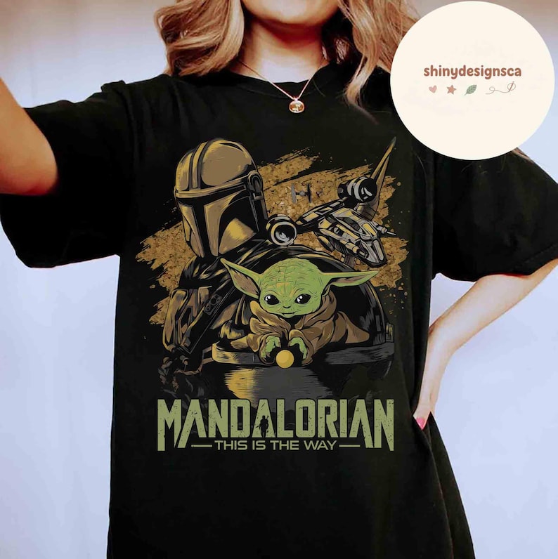 Puede incluir: Camiseta negra con un gr&aacute;fico de The Mandalorian y Grogu, una nave espacial y el texto "MANDALORIAN - THIS IS THE WAY" en verde y dorado. El dise&ntilde;o incluye una figura con casco y un personaje alien&iacute;gena verde.