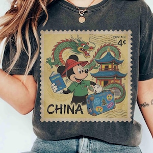 May include: Dark grey t-shirt with a vintage-style graphic of Mickey Mouse in China. The design includes a dragon, pagoda, and the word "CHINA" in a postage stamp design. Mickey is holding a passport and suitcase.