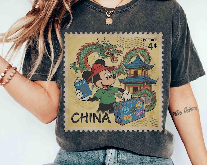 May include: Dark grey t-shirt with a vintage-style graphic of Mickey Mouse in China. The design includes a dragon, pagoda, and the word "CHINA" in a postage stamp design. Mickey is holding a passport and suitcase.
