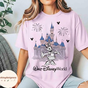 May include: A light pink t-shirt featuring a graphic of Daisy Duck in front of Cinderella's castle, fireworks, and Mickey Mouse silhouettes. The text "Walt Disney World" is printed below the graphic.