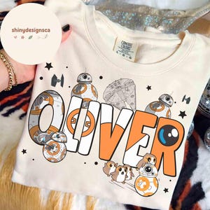 Può includere: Felpa color crema con il nome "OLIVER" in lettere arancioni e bianche, con illustrazioni a tema Star Wars. Il design include droidi BB-8, un Millennium Falcon e altri personaggi. La maglietta è del marchio Comfort Colors.