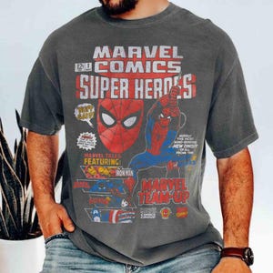 May include: A charcoal gray short-sleeve t-shirt featuring a vintage Marvel Comics graphic. The print displays the text Marvel Comics Super Heroes, a Spider-Man mask, a Spider-Man figure, and smaller panels showing Iron Man and Captain America.