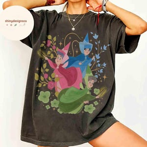 May include: Dark gray t-shirt featuring a colorful illustration of three fairy godmothers from a classic animated film. The design includes floral elements and stars, with the text "shinydesignsca" in the upper left corner.