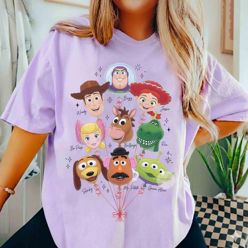 Op de afbeelding: Een lichtpaars t-shirt met een grafische print van diverse Toy Story-personages ontworpen als ballonnen. De personages zijn onder andere Woody, Buzz Lightyear, Jessie, Bo Peep, Bullseye, Rex, Slinky, Mr. Potato Head en een groene Alien.