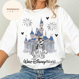 May include: White t-shirt featuring a graphic of Pluto in front of Cinderella's castle at Walt Disney World. The design includes fireworks and Mickey Mouse silhouettes. The text "Walt Disney World" is printed below the graphic.