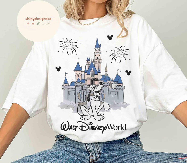 May include: White t-shirt featuring a graphic of Pluto in front of Cinderella's castle at Walt Disney World. The design includes fireworks and Mickey Mouse silhouettes. The text "Walt Disney World" is printed below the graphic.