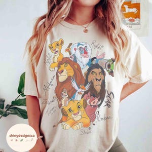Op de afbeelding: Crèmekleurig T-shirt met een kleurrijke afbeelding van personages uit The Lion King, waaronder Simba, Mufasa en Scar. De namen van de personages zijn in een scriptlettertype geschreven. Het shirt heeft een relaxte pasvorm.