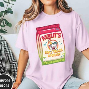 Puede incluir: Camiseta rosa claro con un gr&aacute;fico de una bolsa de snacks con el texto "Maui's" y "Boat Snack Upgrade". El gr&aacute;fico incluye personajes de dibujos animados. La camiseta es de estilo comfort colors.