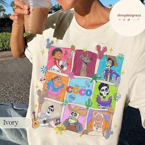 May include: Ivory-colored t-shirt featuring a colorful graphic of characters from the Disney Pixar movie "Coco." The design includes various characters in a grid layout, with the movie's title prominently displayed. The shirt has a crew neck.