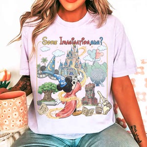 Op de afbeelding: Lichtpaars T-shirt met een kleurrijke afbeelding van Mickey Mouse als tovenaar, een kasteel en de tekst "Some Imagination, Huh?". Het ontwerp bevat wolken, palmbomen en andere themaparkelementen.