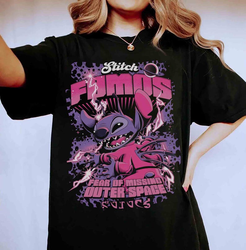 May include: Black t-shirt featuring a graphic of Stitch from Lilo & Stitch. The design includes the words "Stitch Famous" and "Fear of Missing Outer Space". The artwork is in shades of purple, pink, and white, with lightning bolt accents.