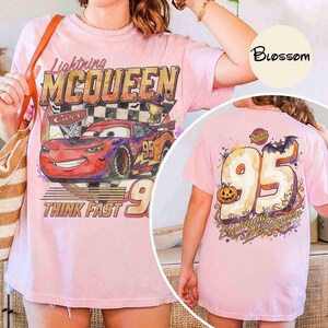 May include: A light pink short-sleeve t-shirt featuring a graphic of the red race car Lightning McQueen with the text Lightning MCQUEEN and THINK FAST. The back of the shirt displays a large number 95 with a pumpkin and a bat graphic.