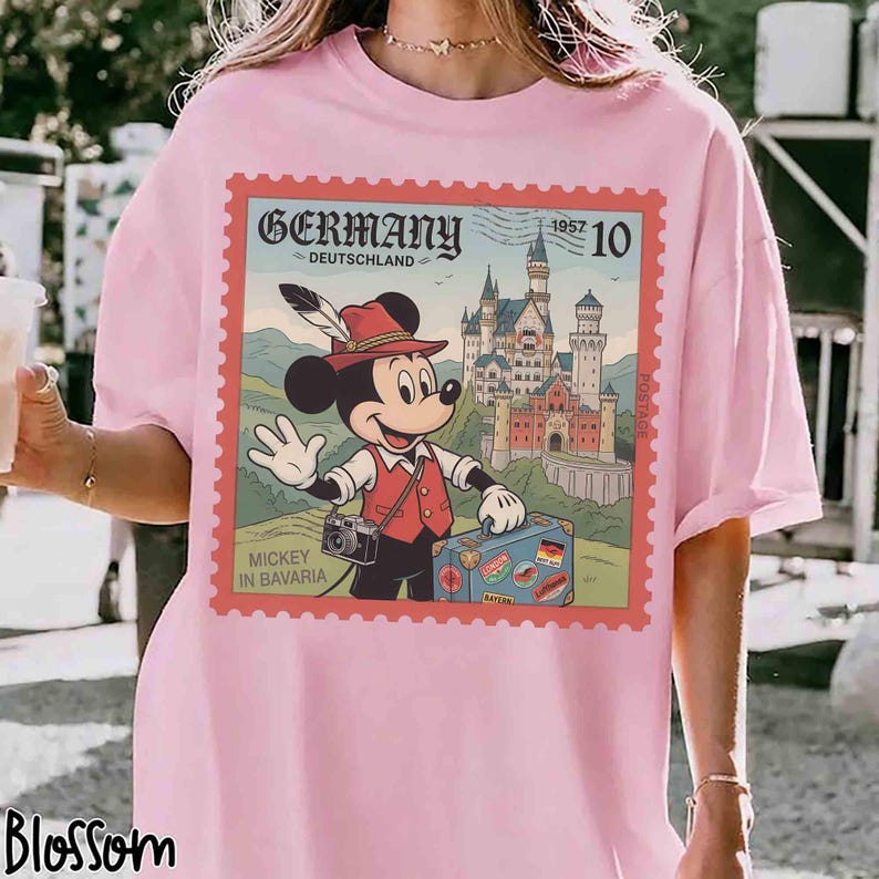May include: Pale pink t-shirt with a postage stamp design. The stamp shows Mickey Mouse in Bavarian clothing, a castle, and the word "Germany". The stamp is framed in red with the text "Mickey in Bavaria".