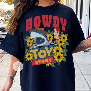 Puede incluir: Camiseta azul marino con un dise&ntilde;o gr&aacute;fico que presenta un tibur&oacute;n con sombrero de vaquero, Woody de Toy Story y las palabras "HOWDY" y "TOY STORY". El dise&ntilde;o est&aacute; rodeado de girasoles amarillos.