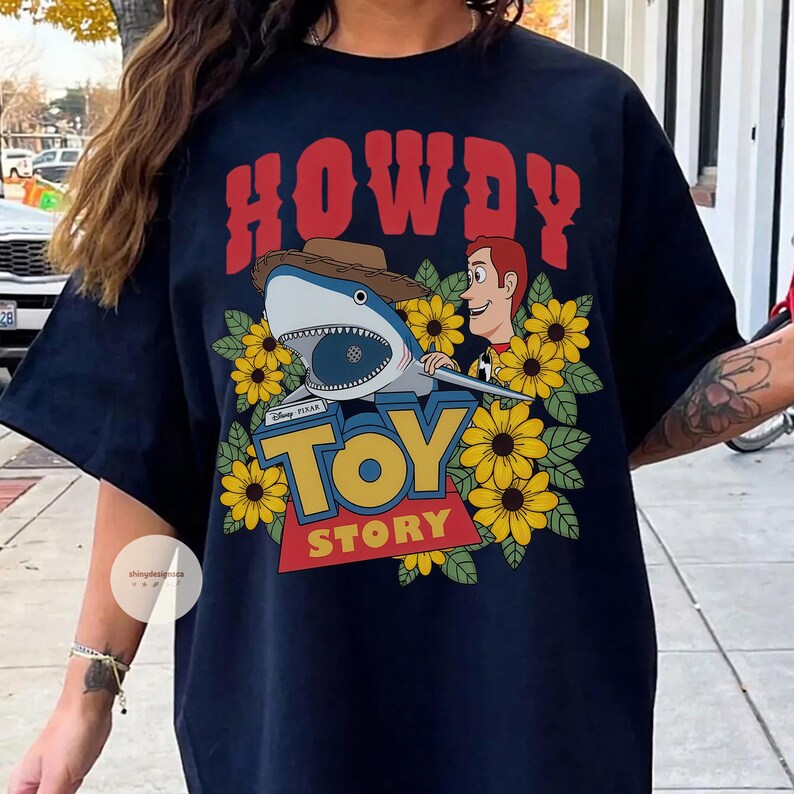 Puede incluir: Camiseta azul marino con un dise&ntilde;o gr&aacute;fico que presenta un tibur&oacute;n con sombrero de vaquero, Woody de Toy Story y las palabras "HOWDY" y "TOY STORY". El dise&ntilde;o est&aacute; rodeado de girasoles amarillos.