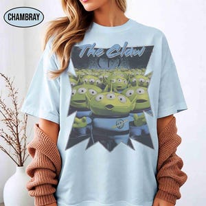 K&ouml;nnte beinhalten: Hellblaues T-Shirt mit einer Grafik der Aliens aus dem Film Toy Story. Die Grafik enth&auml;lt den Text "The Claw" und eine Gruppe gr&uuml;ner, dre&auml;ugiger Aliens. Das T-Shirt hat kurze &Auml;rmel und einen Rundhalsausschnitt.