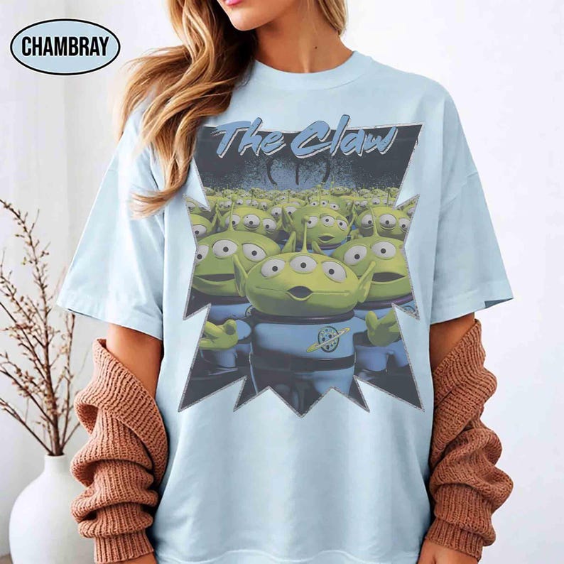 K&ouml;nnte beinhalten: Hellblaues T-Shirt mit einer Grafik der Aliens aus dem Film Toy Story. Die Grafik enth&auml;lt den Text "The Claw" und eine Gruppe gr&uuml;ner, dre&auml;ugiger Aliens. Das T-Shirt hat kurze &Auml;rmel und einen Rundhalsausschnitt.