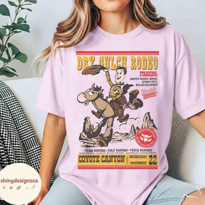 Puede incluir: Una camiseta rosa claro con un gr&aacute;fico retro que anuncia el "Dry Gulch Rodeo". El dise&ntilde;o muestra un vaquero de dibujos animados a caballo, con el texto "Starring: Sheriff Woody, Jessie, Stinky Pete, Bullseye & More!!"