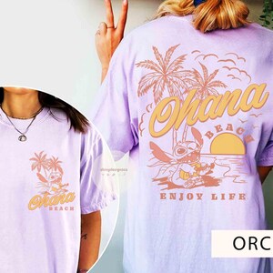 May include: Orchid-colored t-shirt featuring a graphic of a cartoon character on a beach with palm trees and the words "Ohana Beach" and "Enjoy Life". The design is repeated on the front and back of the shirt.