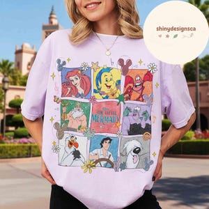 May include: A light purple t-shirt featuring a colorful graphic of characters from Disney's "The Little Mermaid." The design includes Ariel, Flounder, Sebastian, King Triton, Ursula, and Prince Eric. The shirt has a crew neck and short sleeves.