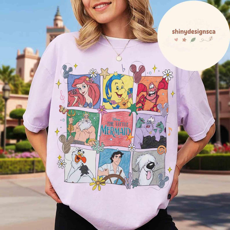 May include: A light purple t-shirt featuring a colorful graphic of characters from Disney's "The Little Mermaid." The design includes Ariel, Flounder, Sebastian, King Triton, Ursula, and Prince Eric. The shirt has a crew neck and short sleeves.